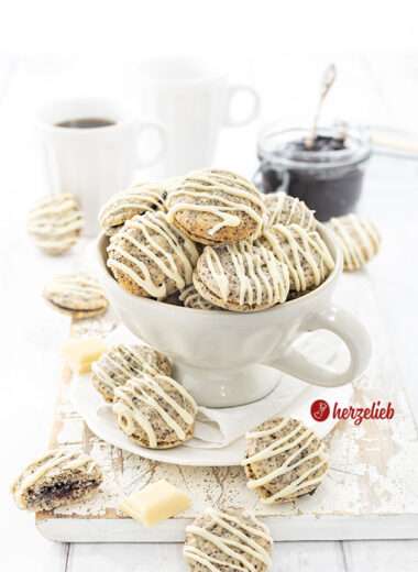 Eine große Tasse, gefüllt mit gefüllten Mohnplätzchen nach einem Germknödel Plätzchen Rezept von herzelieb. Die Plätzchen sind mit weißer Kuvertüre verziert. . Im Hintergrund ein Glas mit Pflaumenmus und zwei Becher Kaffee.