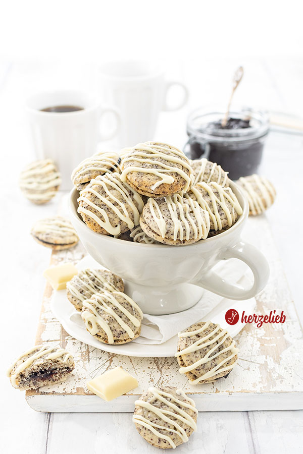 Eine große Tasse, gefüllt mit gefüllten Mohnplätzchen nach einem Germknödel Plätzchen Rezept von herzelieb. Die Plätzchen sind mit weißer Kuvertüre verziert. . Im Hintergrund ein Glas mit Pflaumenmus und zwei Becher Kaffee. 