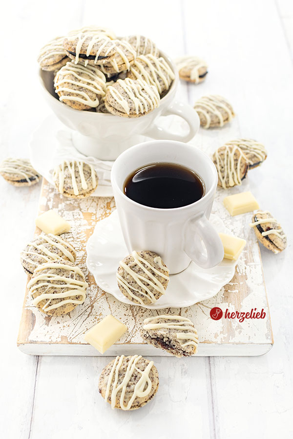 Gefülllte Mohnplätzchen nach einem Germknödel Plätzchen Rezept von herzelieb. Arrangiert zu einer Tasse Kaffee. Im Hintergrund eine große Tasse mit den Plätzchen, die mit weißer Kuvertüre serviert sind.