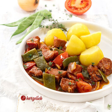 Saftiges Kasseler Gulasch mit Bohnen, Tomaten, Kasseler Nacken und Zwiebeln in dunkler Soße. Serviert mit Katoffeln. Im Hintergrund Stangenbohnen, eine halbe Tomate und zwei Zwiebeln.