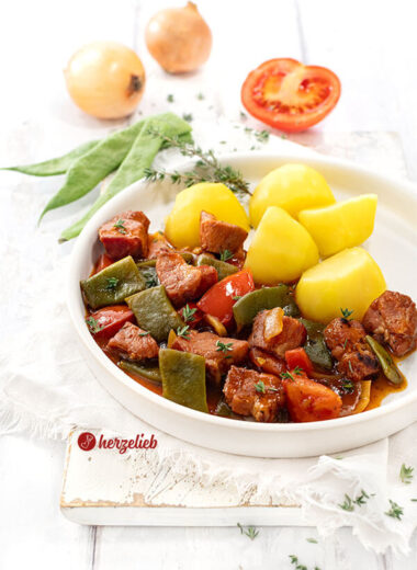 Saftiges Kasseler Gulasch mit Bohnen, Tomaten, Kasseler Nacken und Zwiebeln in dunkler Soße. Serviert mit Katoffeln. Im Hintergrund Stangenbohnen, eine halbe Tomate und zwei Zwiebeln.