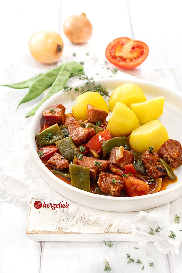 Saftiges Kasseler Gulasch mit Bohnen, Tomaten, Kasseler Nacken und Zwiebeln in dunkler Soße. Serviert mit Katoffeln. Im Hintergrund Stangenbohnen, eine halbe Tomate und zwei Zwiebeln.