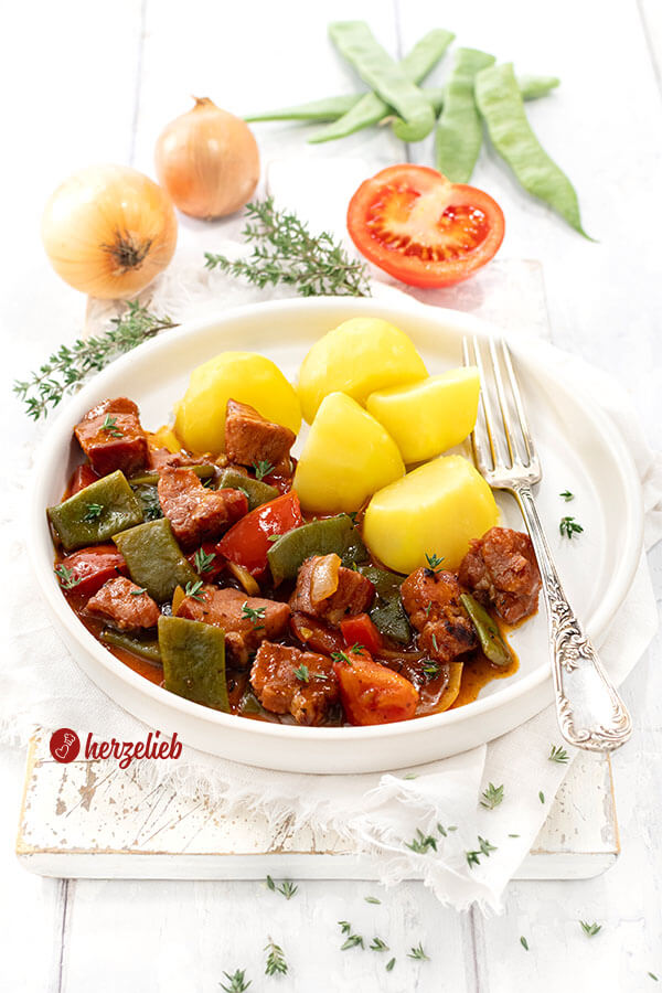 Kasseler Gulasch mit Kartoffeln. Im Gericht sieht man Tomaten, Kasseler Nacken Würfel, Zwiebeln und Bohnen in dunkler Soße.