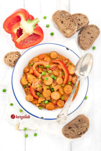 Eine weiße Emailleschüssel mit einem Würstcheneintopf nach einem Piratgryde Rezept aus Dänemark. Im Eintopf sieht man Würstchenscheiben, Paprika und Erbsen in einer tomatigen Soße. Serviert mit Brot, auf dem Schüsselrand ein Löffel