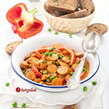 Ein Würstcheneintopf in weißer Emailleschüssel nach einem Piratgryde Rezept von herzelieb. Im Eintopf sind Würstchenscheiben, Paprikastücke und Erbsen in cremiger Tomatensoße. Im Hintergrund ein Korb mit Brot, eine halbe rote Paprikaschote. Auf dem Schüsselrand ein Silberlöffel.