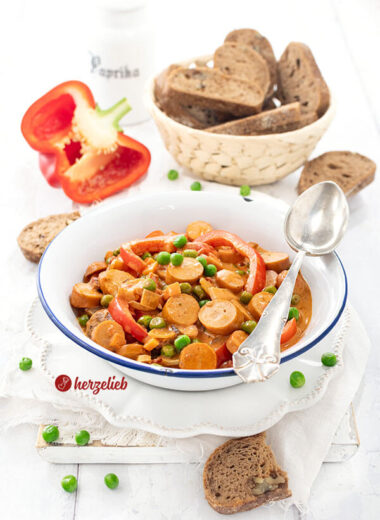 Ein Würstcheneintopf in weißer Emailleschüssel nach einem Piratgryde Rezept von herzelieb. Im Eintopf sind Würstchenscheiben, Paprikastücke und Erbsen in cremiger Tomatensoße. Im Hintergrund ein Korb mit Brot, eine halbe rote Paprikaschote. Auf dem Schüsselrand ein Silberlöffel.
