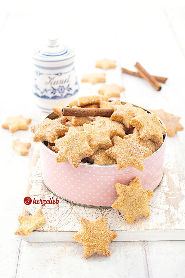 Knusprige Plätzchen in Sternform mit einer Zimt-Zucker-Kruste nach einem Rahmplätzchen Rezept von herzelieb. Angerichtet in einer rosafarbenen Dose mit weißen Punkten. Dekoriert mit Zimtstangen, im HIntergrund ein weißes Zimtgefäß mit blauem Schmuckdekor.