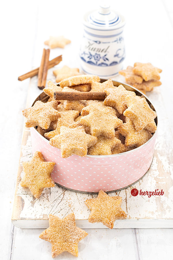 In einer rosa Dose sind Plätzchen nach einem Rahmplätzchen Rezept von herzelieb gestapelt. Die Kekse sind goldbraun gebacken und haben eine Zuckerkruste. Dekoriert mit Zimtstangen