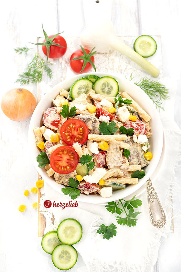 Bunter Gyros Salat in einer weißen Schüssel. Der herzhafte Nudelsalat ist in einet weißen Schüssel angerichtet und rundherum dekoriert mit Gemüse und Kräutern. 