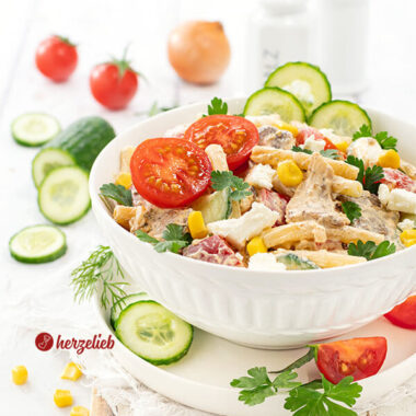 Gyros Salat in einer weißen Schale. Der Nudesalat ist dekoriert mit Gurken, Tomaten, glatter Petersilie und Mais. Im Hintergrund Gemüse.