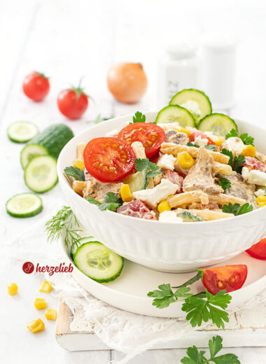 Gyros Salat in einer weißen Schale. Der Nudesalat ist dekoriert mit Gurken, Tomaten, glatter Petersilie und Mais. Im Hintergrund Gemüse.