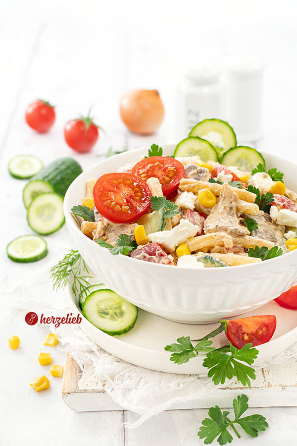 Gyros Salat in einer weißen Schale. Der Nudesalat ist dekoriert mit Gurken, Tomaten, glatter Petersilie und Mais. Im Hintergrund Gemüse.