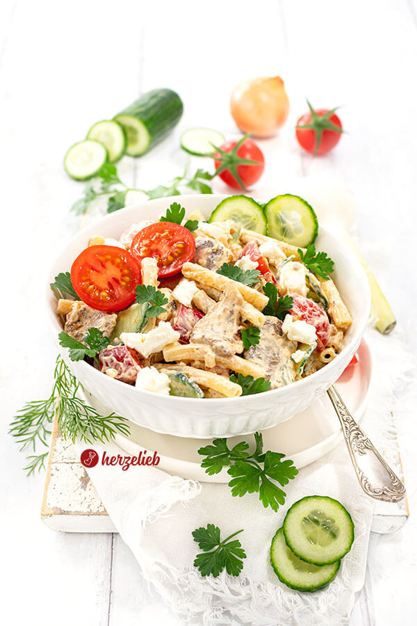Nudelsalat nach einem Gyrossalat Rezept von herzelieb. Angerichtet in einer weißen Schale, dekoriert mit Gurken, tomaten und glatter Petersilie. Auf dem Unterteller eine Gabel, im Vordergrund Kräuter, Im Hintergrund Gemüse