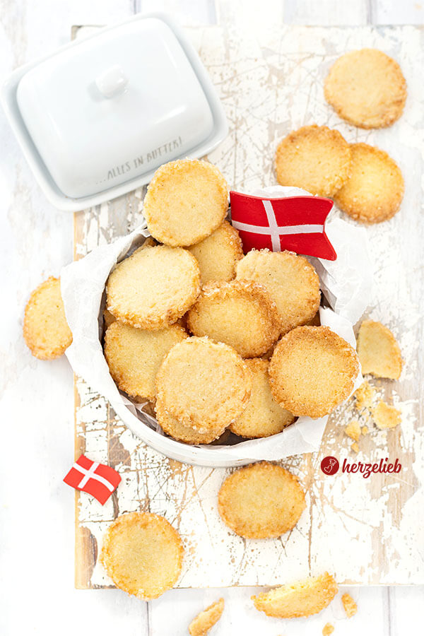 Dänische Plätzchen mit knusprigen Zuckerrand nach einem Håkonskager Rezept aus Dänemark von herzelieb. Angerichtet in einer Dose und dekoriert mit zwei Dannebrog. Im Hintergrund ein Vanillegefäß und eine Butterglocke.