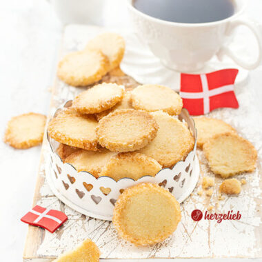 Hellbraune Plätzchen aus Dänemark mit Zuckerrand nach einem Håkonskager Rezept von herzelieb. Angerichtet in einer Dose und dekoriert mit zwei Dannebrog. Im Hintergrund ein Vanillegefäß und eine Butterglocke.