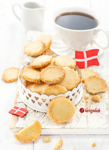 Hellbraune Plätzchen aus Dänemark mit Zuckerrand nach einem Håkonskager Rezept von herzelieb. Angerichtet in einer Dose und dekoriert mit zwei Dannebrog. Im Hintergrund ein Vanillegefäß und eine Butterglocke.