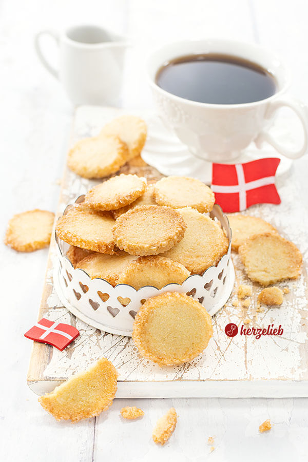 Hellbraune Plätzchen aus Dänemark mit Zuckerrand nach einem Håkonskager Rezept von herzelieb. Angerichtet in einer Dose und dekoriert mit zwei Dannebrog. Im Hintergrund ein Vanillegefäß und eine Butterglocke.