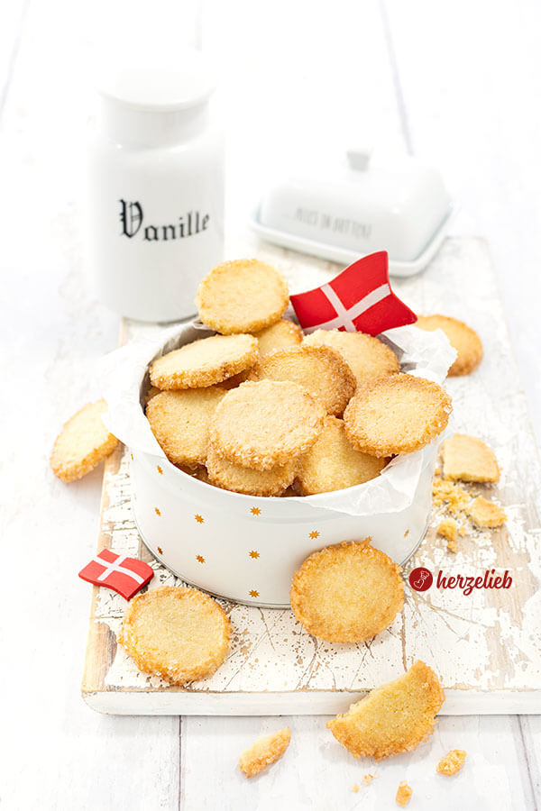 Leckere, hellbraune Plätzchen mit Zuckerrand nach einem Håkonskager Rezept von herzelieb. Angerichtet in einer Dose und dekoriert mit zwei Dannebrog. Im hintergrnd ein Vanillegefäß und eine Butterglocke