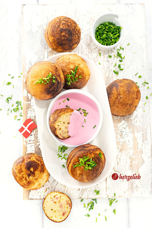 Einige goldbraun gebackene, herzhafte Æbleskiver auf einer weißen, ovalen Porzellanplatte. Serviert mit einem Schälchen Rote Bete Frischkäse. Die Gebäckbällchen sind mit gehacktem Schnittlauch dekoriert.