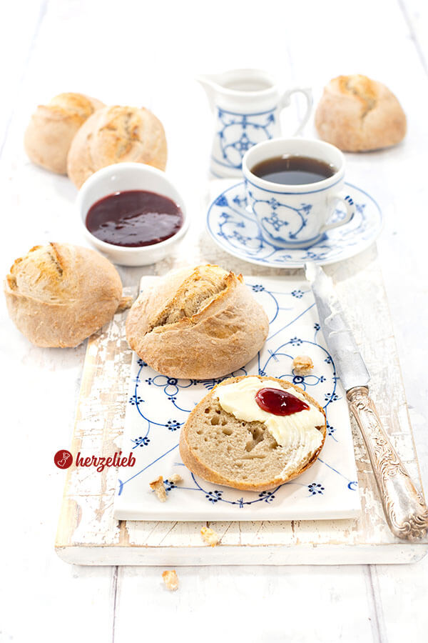 Ein Frühstückstisch mit frisch gebackenen Brötchen nach einem Ohne-Kneten-Brötchen Rezept von herzelieb. Angericht auf blauweiß gemustertem Geschirr. Im Hintergrund ein Schälchen Marmelade und eine Tasse Kaffee.