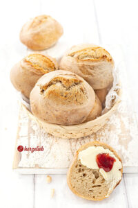 Ein Körbchen mit frisch gebackenen Brötchen nach einem Ohne-Kneten-Brötchen Rezept von herzelieb. Vor dem Korb liegt eine bestrichene Brötchenhälfte.