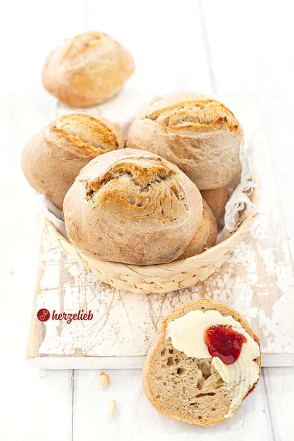 Ein Körbchen mit frisch gebackenen Brötchen nach einem Ohne-Kneten-Brötchen Rezept von herzelieb. Vor dem Korb liegt eine bestrichene Brötchenhälfte.
