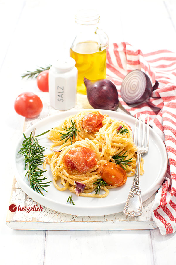 Ein weißer Teller mit hohem Rand und einem Nudelgericht nach einem Feta-Tomaten-Pasta Rezept von herzelieb. Im Hintergrund Tomaten, ein Salzstreuer, eine Flasche Olivenöl und Tomaten. Das Pastagericht ist dekoriert mit Rosmarinzweigen