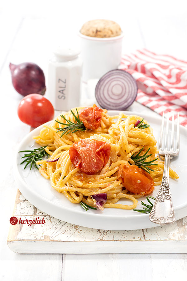 Ein Teller mit einem Nudelgericht nach einem Feta-Tomaten-Pasta Rezept von herzelieb. Angerichtet auf einem weißen Teller mit Tomaten, dekoriert mit Rosmarin. Im Hintergrund rote Zwiebeln, eine Tomate ein Salzstreuer und ein rot-weiß gestreiftes Tuch.