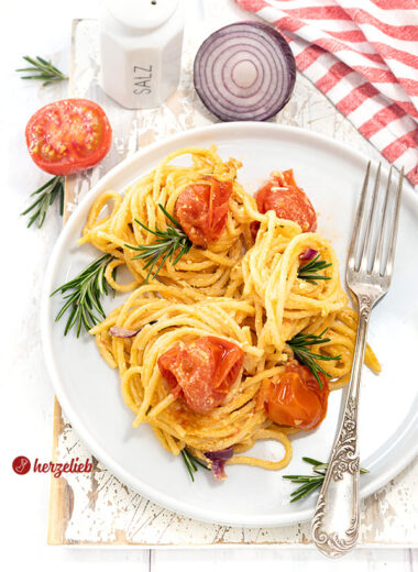 Leckeres Nudelgericht nach einem Feta-Tomaten-Pasta Rezept von herzelieb. Angerichtet auf einem weißen Teller, dekoriert mit Rosmarin. Im Hintergrund Tomate, rote Zwiebel und ein Salzstreuer. Auf dem Tellerrand liegt eine silberne Gabel.