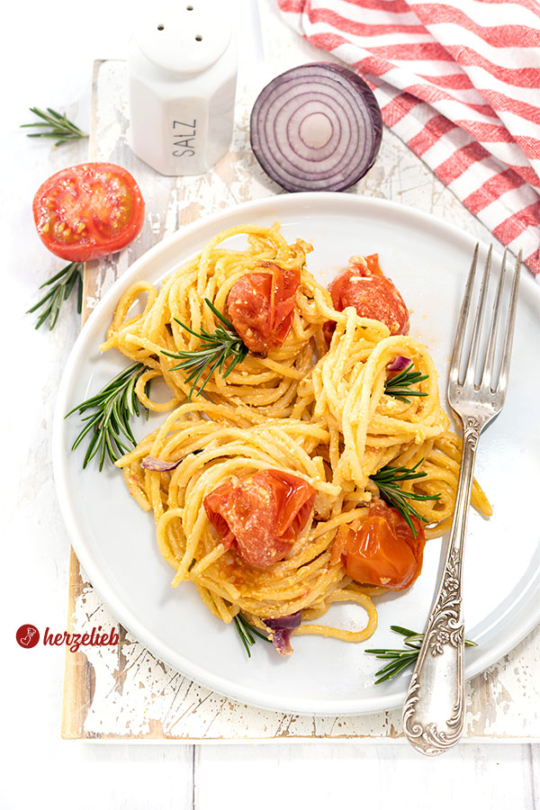Leckeres Nudelgericht nach einem Feta-Tomaten-Pasta Rezept von herzelieb. Angerichtet auf einem weißen Teller, dekoriert mit Rosmarin. Im Hintergrund Tomate, rote Zwiebel und ein Salzstreuer. Auf dem Tellerrand liegt eine silberne Gabel.