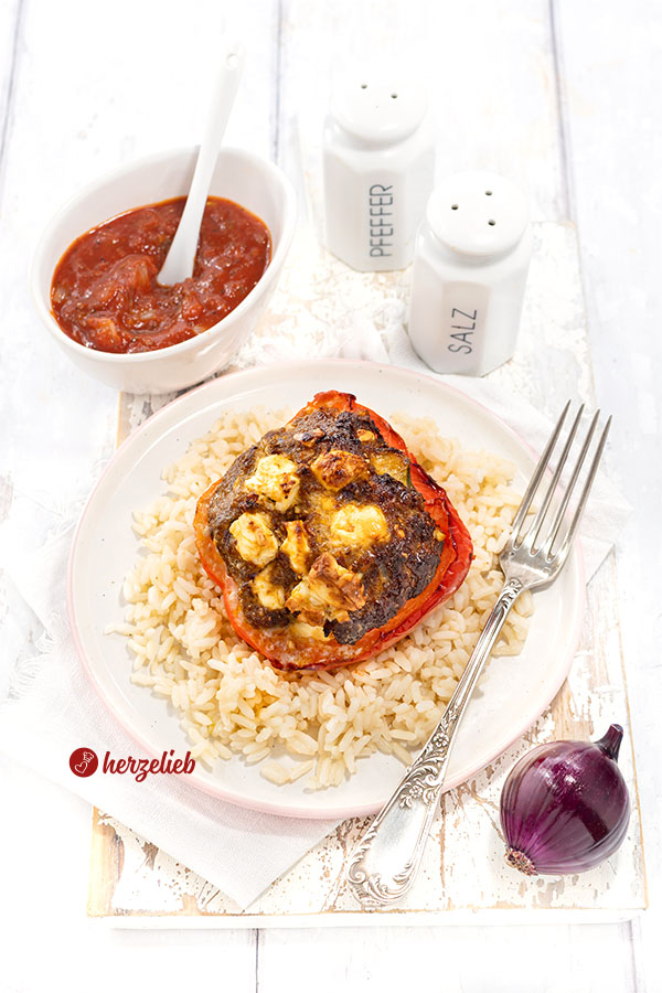 Paprika, Hack, Feta – die Kombination zum gefüllte Paprikaschoten Rezept von herzelieb. Die gefüllte rote Paprika liegt auf einem Reisbett. Am Tellerrand eine Gabel, im Hintergrund eine Schale Tomatensoße sowie ein Pfeffer- und ein Salzstreuer.