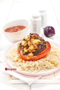 Eine rote Paprika mit einer Füllung aus Hack und Feta nach einem gefüllte Paprikaschoten Rezept mit Fetavon herzelieb. Angerichtet auf Reis. Im Hintergrund Tomatensauce, eine rote Zwiebel und ein Pfeffer- sowie ein Salzstreuer.