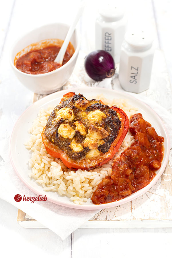 Eine mit Hackfleisch und Feta gefüllte Paprika nach einem gefüllte Paprikaschoten Rezept von herzelieb. Angerichtet auf Reis mit Tomatensoße. Im Hintergrund eine Schale Sauce, ein Salz- und ein Pfefferstreuer und eine rote Zwiebel.