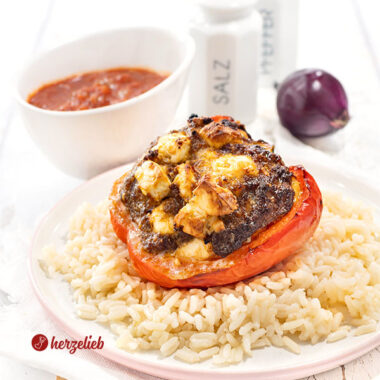 Eine rote Paprika mit einer Füllung aus Hack und Feta nach einem gefüllte Paprikaschoten Rezept mit Fetavon herzelieb. Angerichtet auf Reis. Im Hintergrund Tomatensauce, eine rote Zwiebel und ein Pfeffer- sowie ein Salzstreuer.