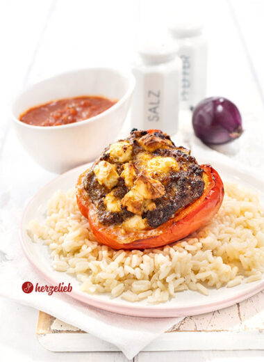Eine rote Paprika mit einer Füllung aus Hack und Feta nach einem gefüllte Paprikaschoten Rezept mit Fetavon herzelieb. Angerichtet auf Reis. Im Hintergrund Tomatensauce, eine rote Zwiebel und ein Pfeffer- sowie ein Salzstreuer.