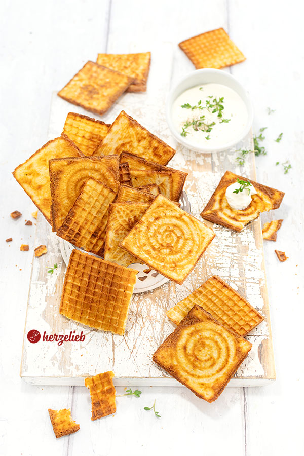 Eine Schale mit Käsewaffeln aus dem Zimtwaffeleisen. Goldgelb gebacken, knusprig, klein und viereckig. Im Hintergrund ein Schälchen Dip, dekoriert mit Kresse