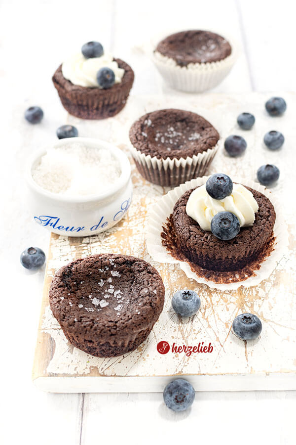 Schokoladenmuffins aus Schweden nach einem Kladdkakemuffins Rezept von herzelieb auf einem weißen Brett. Zwei sind bestreut mit Salzflocken, zwei mit Sahne und Blaubeeren dekoriert.