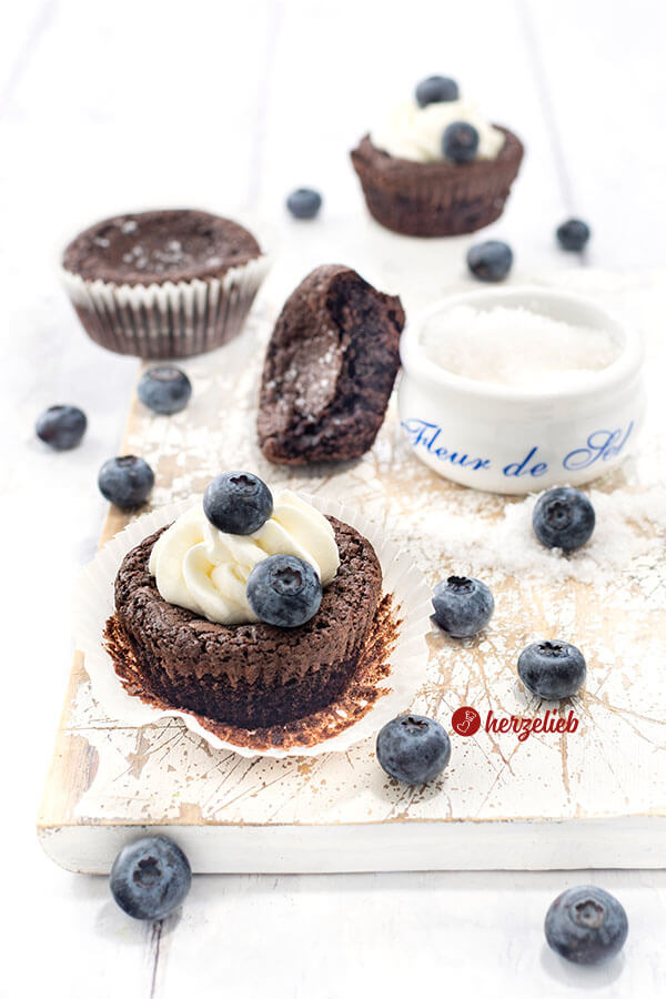 Schwedischen Schokoladenmuffins nach einem Kladdkakemuffins Rezept von herzelieb. Im Vordergrund mit Sahne und Blaubeeren. Dahinter ein Salzgefäß und ein halber Muffin. Im Hintergrund weitere kleine Schokoladenkuchen