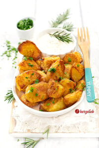 Krosse und goldbraun gebackene Kartoffeln nach einen Kusperkartoffeln Rezept von herzelieb. Die Knoblauchkartoffeln sind mit einem Schälchen Dip auf einer ovalen Porzellanplatte angerichtet. Auf dem Tellerrand liegt eine Holzgabel.
