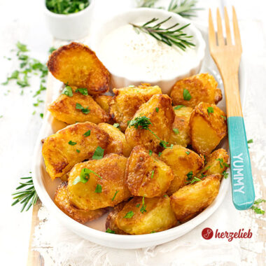 Krosse und goldbraun gebackene Kartoffeln nach einen Kusperkartoffeln Rezept von herzelieb. Die Knoblauchkartoffeln sind mit einem Schälchen Dip auf einer ovalen Porzellanplatte angerichtet. Auf dem Tellerrand liegt eine Holzgabel.