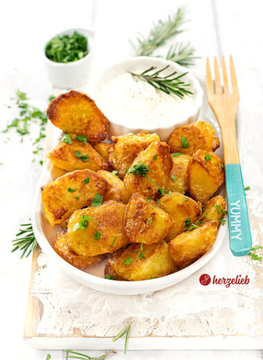 Krosse und goldbraun gebackene Kartoffeln nach einen Kusperkartoffeln Rezept von herzelieb. Die Knoblauchkartoffeln sind mit einem Schälchen Dip auf einer ovalen Porzellanplatte angerichtet. Auf dem Tellerrand liegt eine Holzgabel.