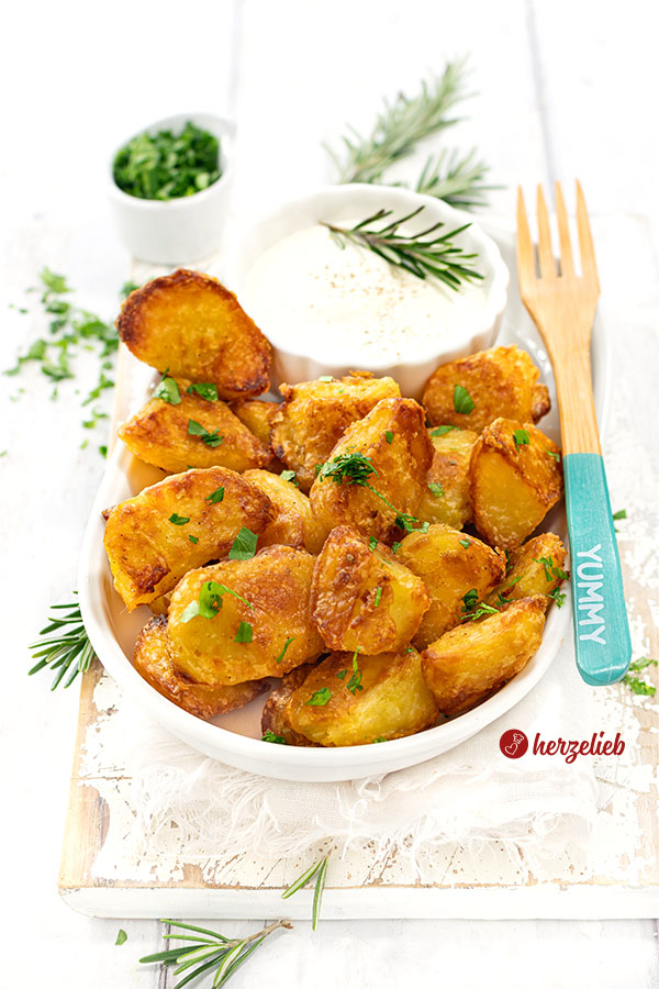 Krosse und goldbraun gebackene Kartoffeln nach einen Kusperkartoffeln Rezept von herzelieb. Die Knoblauchkartoffeln sind mit einem Schälchen Dip auf einer ovalen Porzellanplatte angerichtet. Auf dem Tellerrand liegt eine Holzgabel.