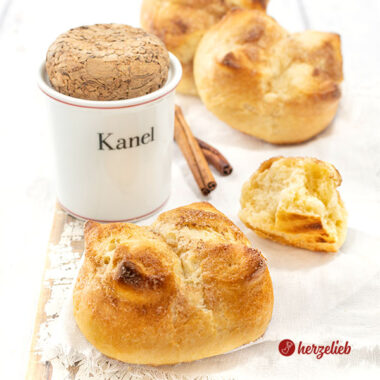 Kronerboller, die weichen Hefebrötchen mit Zimt und Butter auf ein weißen Tuc. Im Hintergrund ein Zimtgefäß, Zimtstangen und weitere Brötchen.
