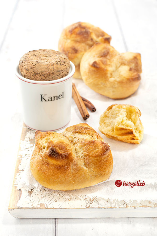 Kronerboller, die weichen Hefebrötchen mit Zimt und Butter auf ein weißen Tuc. Im Hintergrund ein Zimtgefäß, Zimtstangen und weitere Brötchen.