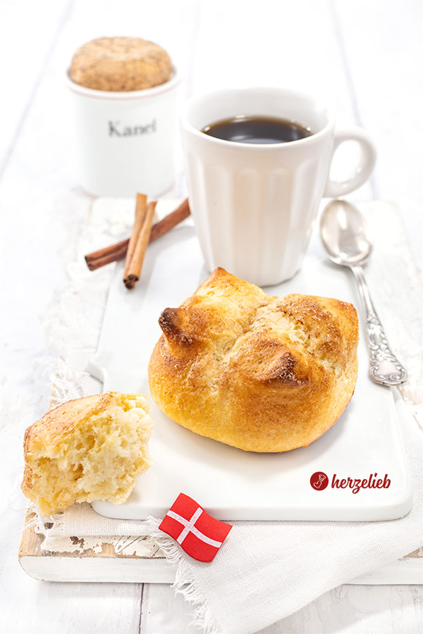 Ein Kronerboller auf einem weißen Porzellan-Frühstücksbrett. Im Hintergrund ein Becher Kaffee Im Vordergrund ein Stück Brötchen.