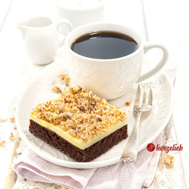 Ein Stück Blechkuchen nach einem Sägespäne Kuchen Rezept von herzelieb mit einem Schokoladenboden, einer Vanillecreme und Kokosknusper auf einem weißen Teller mit einer Tasse Kaffee und einer Kuchengabel auf dem Tellerrand.