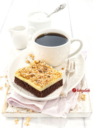 Ein Stück Blechkuchen nach einem Sägespäne Kuchen Rezept von herzelieb mit einem Schokoladenboden, einer Vanillecreme und Kokosknusper auf einem weißen Teller mit einer Tasse Kaffee und einer Kuchengabel auf dem Tellerrand.