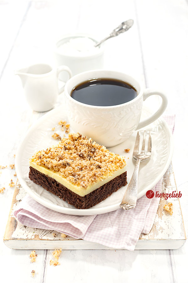 Ein Stück Blechkuchen nach einem Sägespäne Kuchen Rezept von herzelieb mit einem Schokoladenboden, einer Vanillecreme und Kokosknusper auf einem weißen Teller mit einer Tasse Kaffee und einer Kuchengabel auf dem Tellerrand.