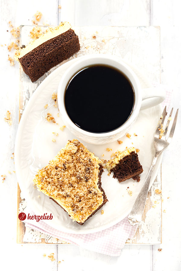 Ein weißer Teller mit einer Tasse Kaffee und einem Stück Blechkuchen nach einem Sägespänekuchen Rezept von herzelieb. Der Kuchen hat 3 Schichten: Schokoboden, Cremeschicht, Kokosknupser.