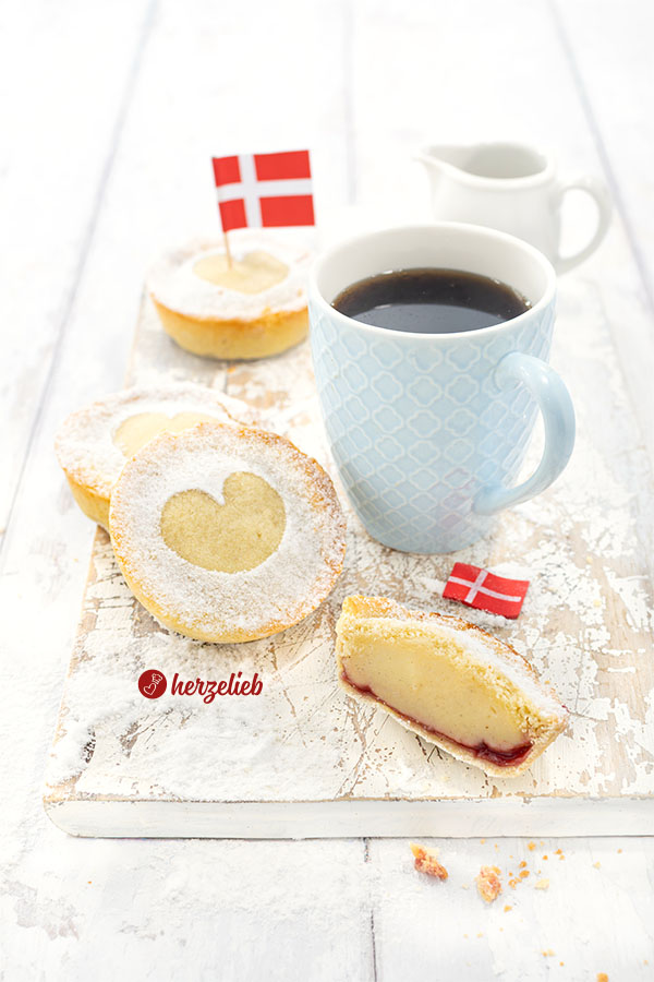 3 kleine Kuchen nach einem Cremelinser Rezept aus Dänemark. Die Kuchen sind mit Puderzucker bestäubt und haben ein Herz in der Mitte. Im Vordergrund ein halbierter Kuchen bei dem man die Cremefüllung und darunter die Marmelade. Im Hintergrund eine Tasse Kaffee.